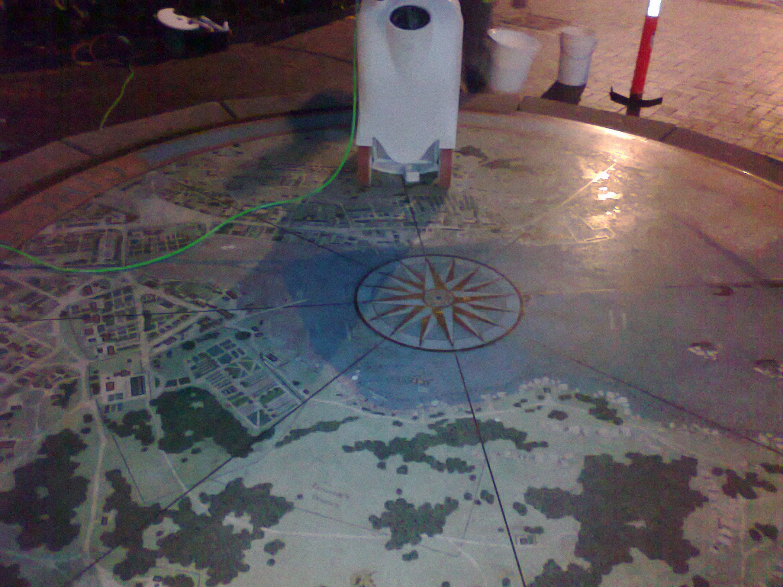 Close-up of the Sydney Cove 1808 Map surface at night, showing the compass rose and inlaid street pattern, with the base of a restoration machine and a power lead resting on the map