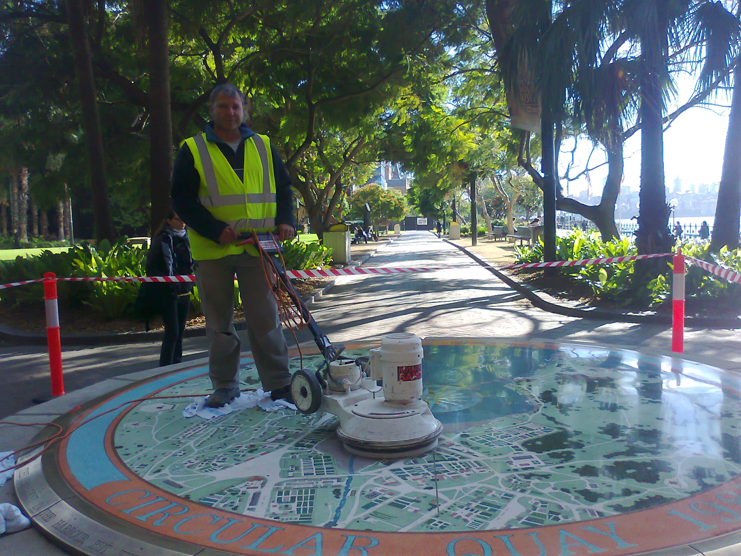 Tony Mastrorillo standing on the Sydney Cove 1808 Map beside a rotary restoration machine, with a tree-lined park path and barrier tape behind him