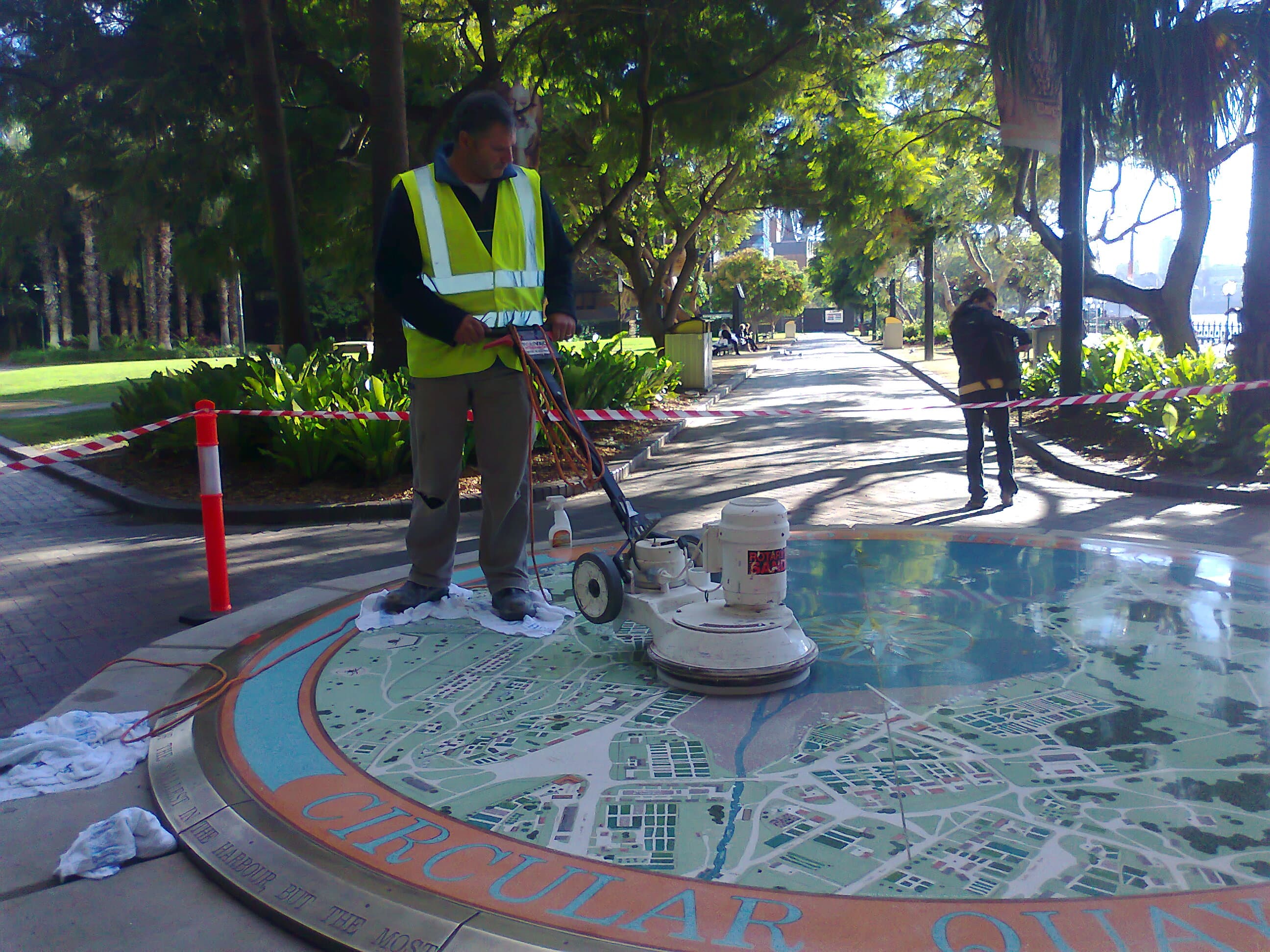 Tony Mastrorillo in a high-visibility vest operating a rotary floor machine on the Sydney Cove 1808 Map, with the Circular Quay border lettering in the foreground and barrier tape around the work area