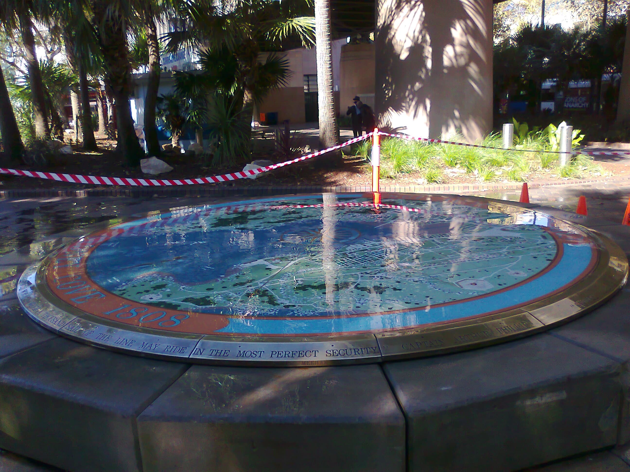 Wide view of the circular Sydney Cove 1808 Map on its raised stone base, showing the Sydney Cove 1808 border lettering, the bronze inscription band and the surrounding park